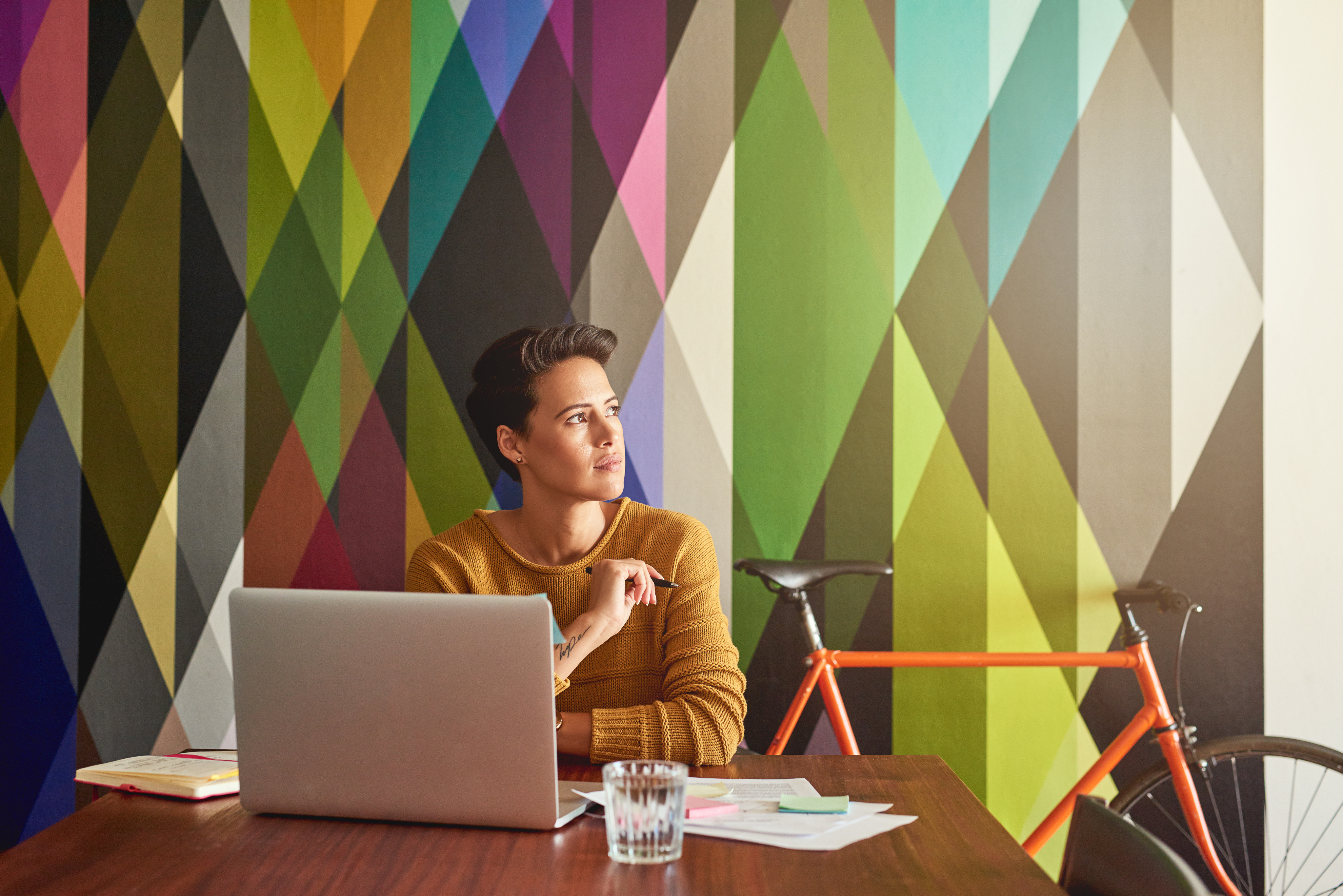 Femme en pull jaune devant un mur graphique multicolore, travaillant sur un MacBook